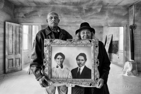 Black and white image of two people stand before a building opening holding a painted portrait.