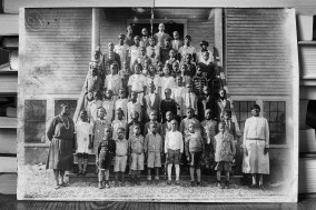 Black and white image of a photograph resting on a table showing students and teachers lined up on a stair case of a school building.