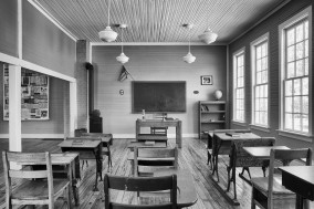 Black and white image of inside a classroom with desks and chairs facing a black chalkboard.