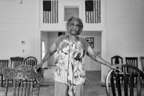 Black and white image of person with arm stretched holding two chairs in front of a building opening.