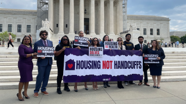 a group of protesters stand outside the Supreme Court, holding a sign that says "Housing not handcuffs"
