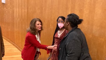 Person greets two others with a handshake before a wood paneled wall.