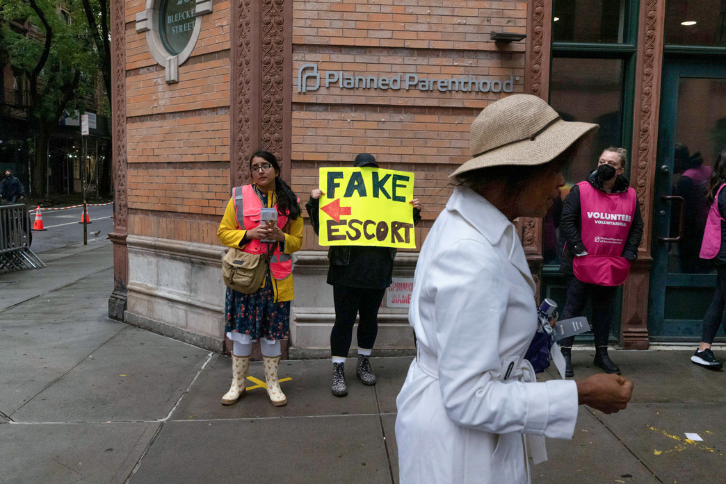 Two people stand along wall with a sign pointing to one person reading "Fake Escort."