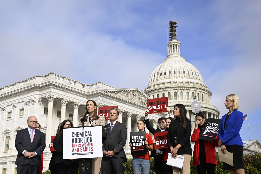 Person at podium is flanked by people holding signs opposing abortion with Capitol building in background.