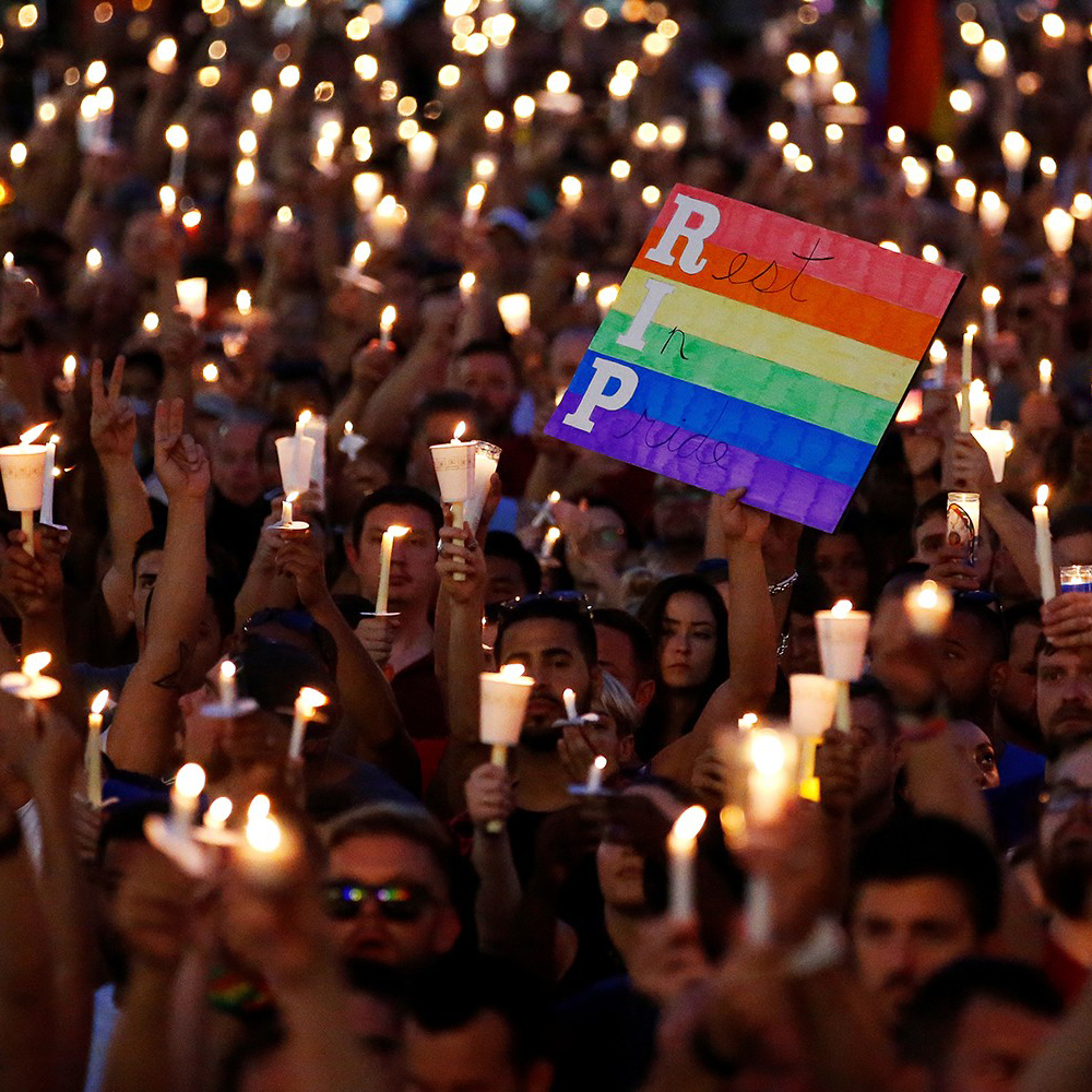 Large vigil of people holding candles with one person holding up a sign with the LGBTQ+ flag with the words "Rest in Pride" written on it.