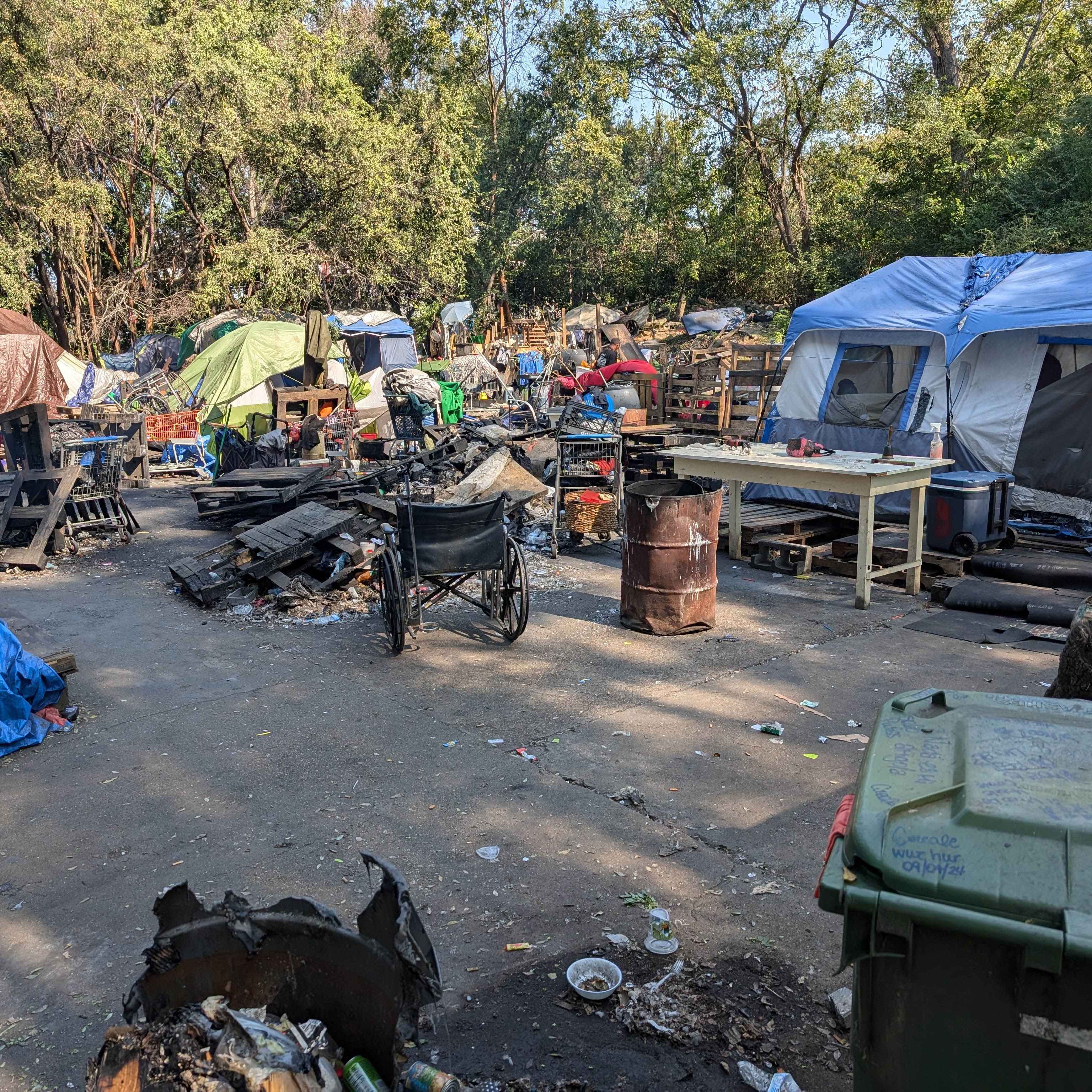 Tents surrounded by trees are grouped together around burned pallets and metal trash barrels. 