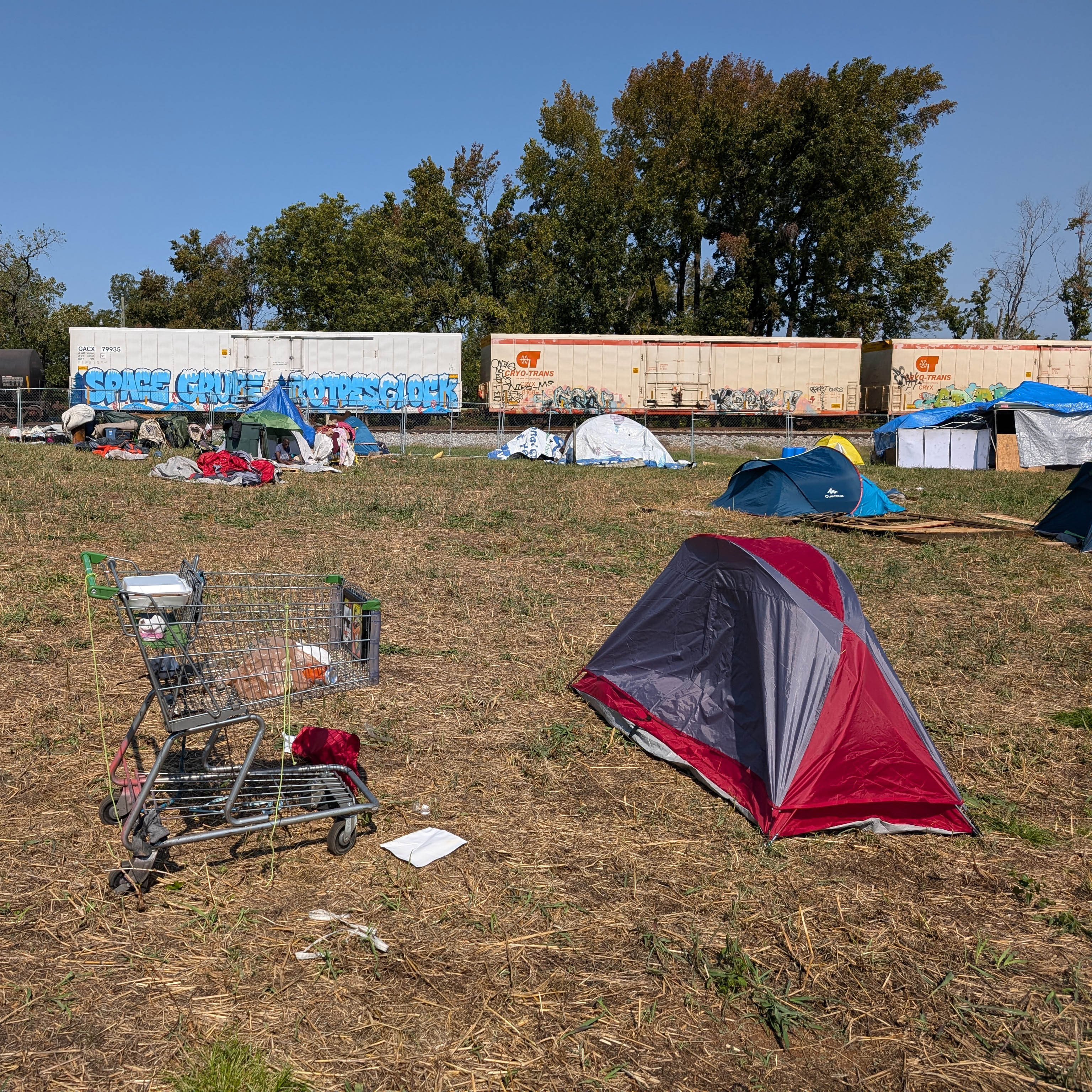 A shopping cart and an erected tent on a field surrounded by a chain-link fence with a train passing in the background