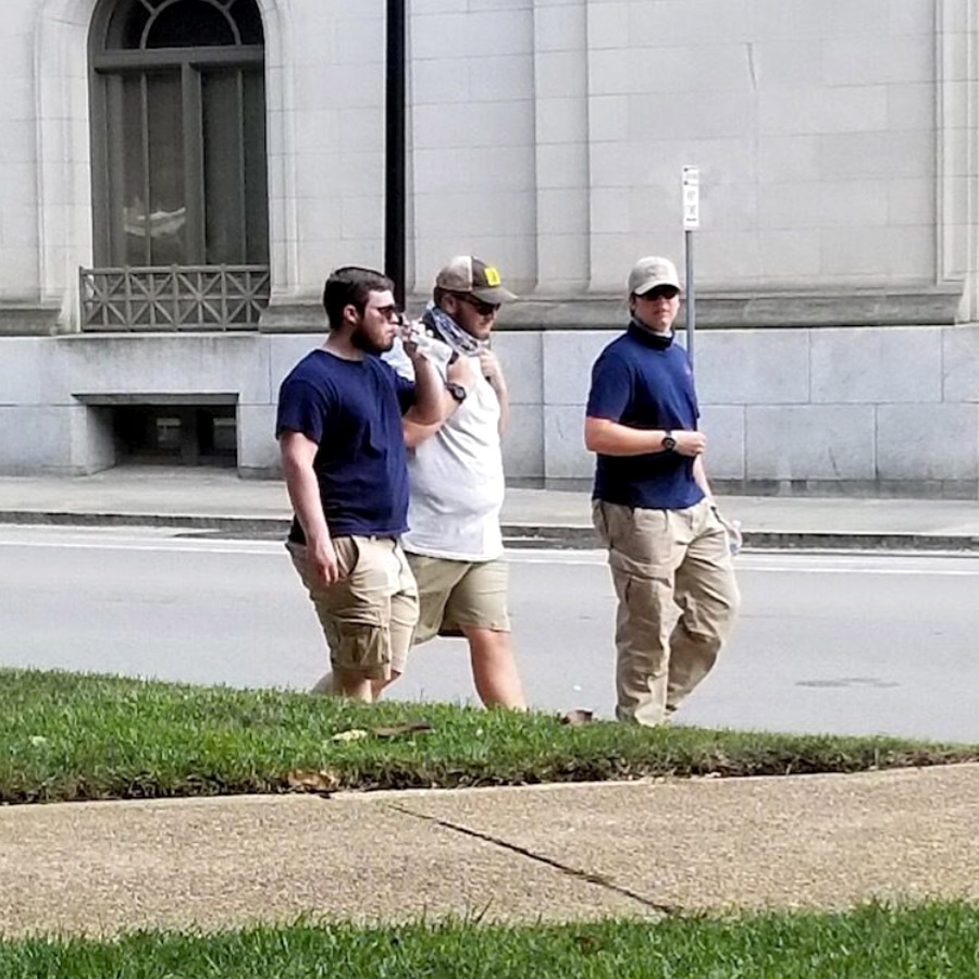 The people in blue shirts and khaki pants flank a third individual dressing in a white shirt as they walk along a city street.