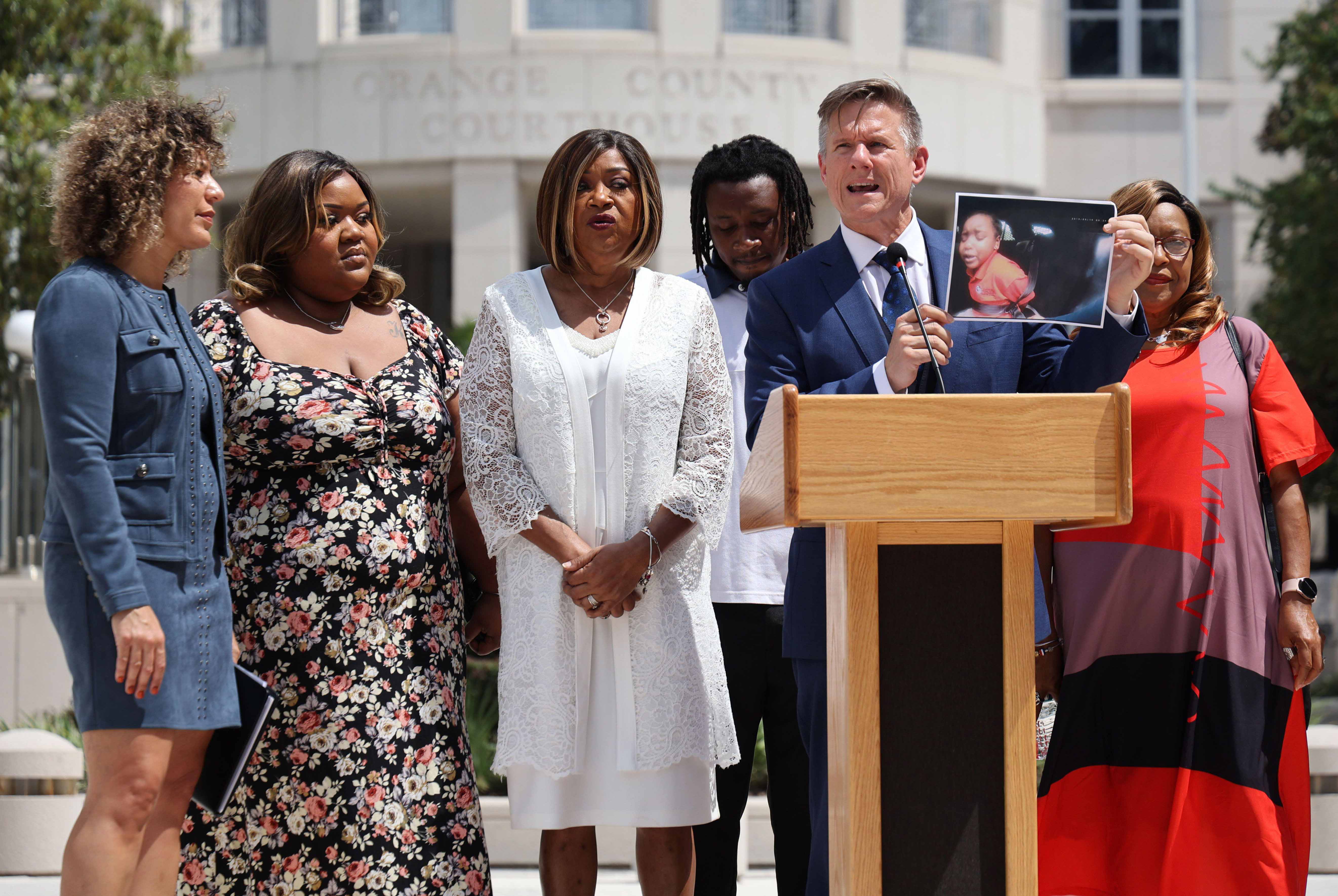 Group of people stand around a podium where a man speaking to a crowd holds up a photo.